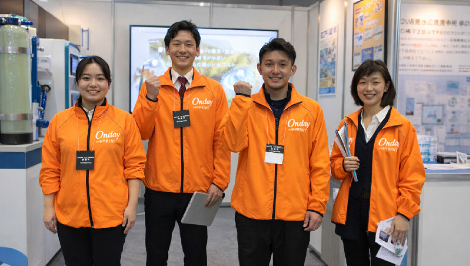企業の展示会でオリジナルプリントのスタッフウェアを着用するチームの様子