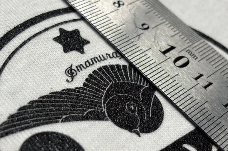 シルクスクリーンプリントの印刷精度をルーラー（定規）で計測している様子。白生地に黒インクで細かなイラストが再現されており、1mm単位の精密な仕上がりを示している