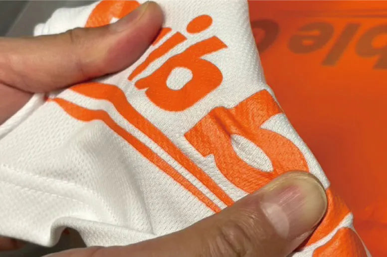 シルクスクリーンプリントの伸縮性を確認している様子。オレンジのロゴがプリントされた白いTシャツを手で引っ張り、生地とインクの密着状態を示している